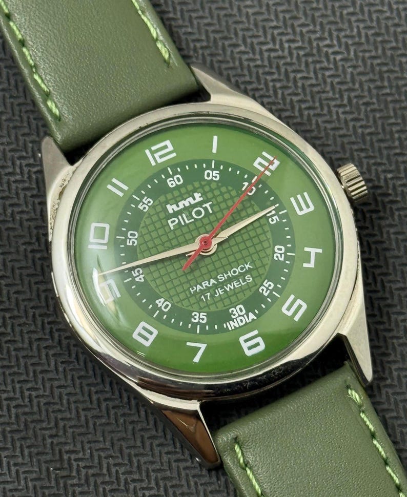 Pode incluir: Um rel&oacute;gio de pulso vintage com mostrador verde e pulseira de couro verde a condizer. O mostrador apresenta algarismos brancos, um ponteiro de segundos vermelho e o texto "hmt PILOT". O rel&oacute;gio tem uma caixa prateada e est&aacute; sobre um fundo escuro.