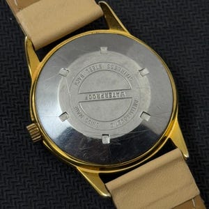 May include: Gold-tone wristwatch with a tan leather band. The back of the watch is stainless steel and says "WATERPROOF", "SWISS MADE", "ANTIMAGNETIC", and "STAINLESS STEEL BACK".