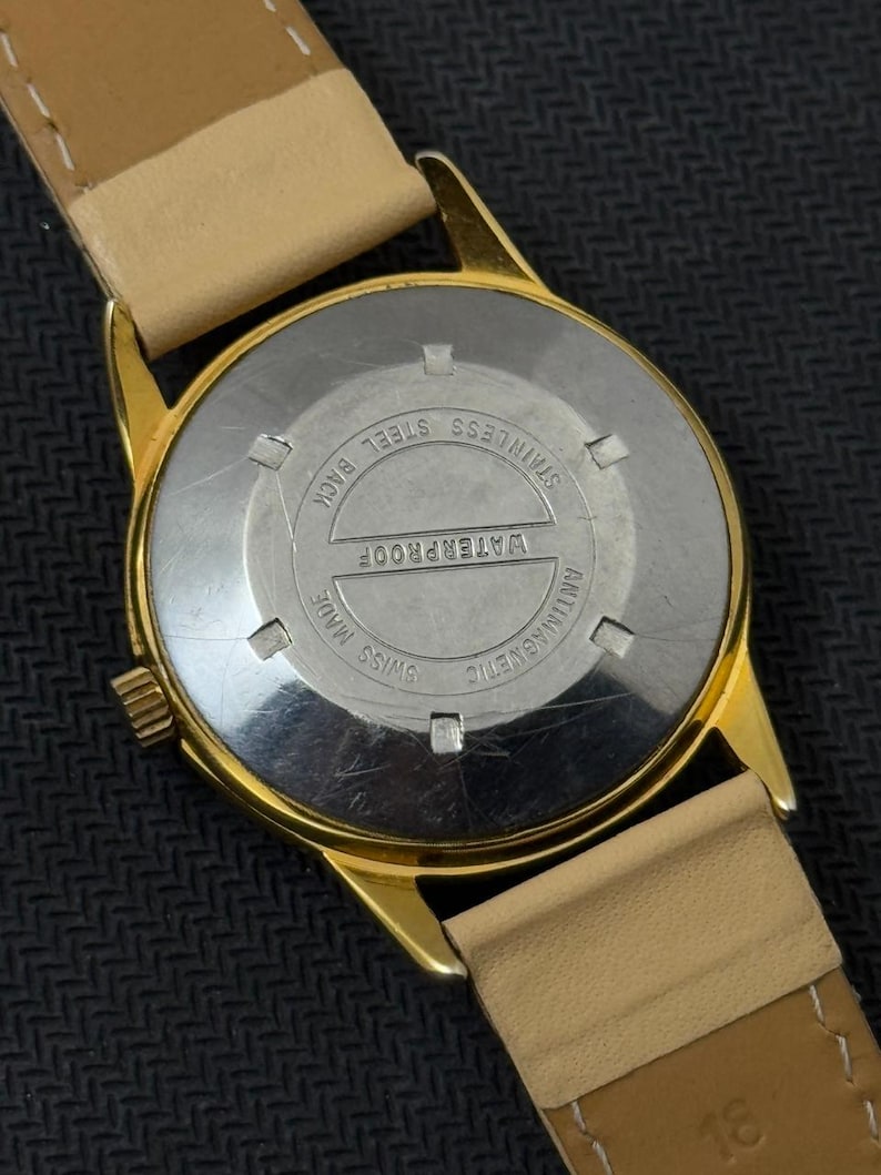 May include: Gold-tone wristwatch with a tan leather band. The back of the watch is stainless steel and says "WATERPROOF", "SWISS MADE", "ANTIMAGNETIC", and "STAINLESS STEEL BACK".