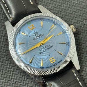 Puede incluir: Un reloj de pulsera Roamer plateado y dorado con una esfera azul y una correa de cuero negro. La esfera del reloj tiene agujas y marcadores dorados, y el texto "AW ROAMER 17 JEWELS SHOCK PROOF" está impreso en la esfera. El reloj está hecho en Suiza.