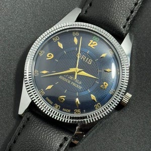 Puede incluir: Un reloj de pulsera vintage con esfera azul oscuro y agujas e indicadores dorados. La esfera muestra la marca "ORIS" y el texto "7 JEWELS SHOCK PROOF". Tiene un bisel plateado y una correa de cuero negro.