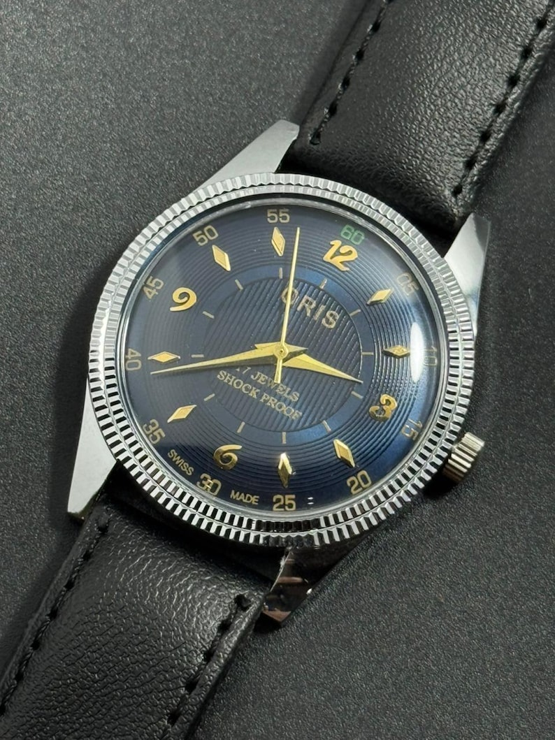Puede incluir: Un reloj de pulsera vintage con esfera azul oscuro y agujas e indicadores dorados. La esfera muestra la marca "ORIS" y el texto "7 JEWELS SHOCK PROOF". Tiene un bisel plateado y una correa de cuero negro.