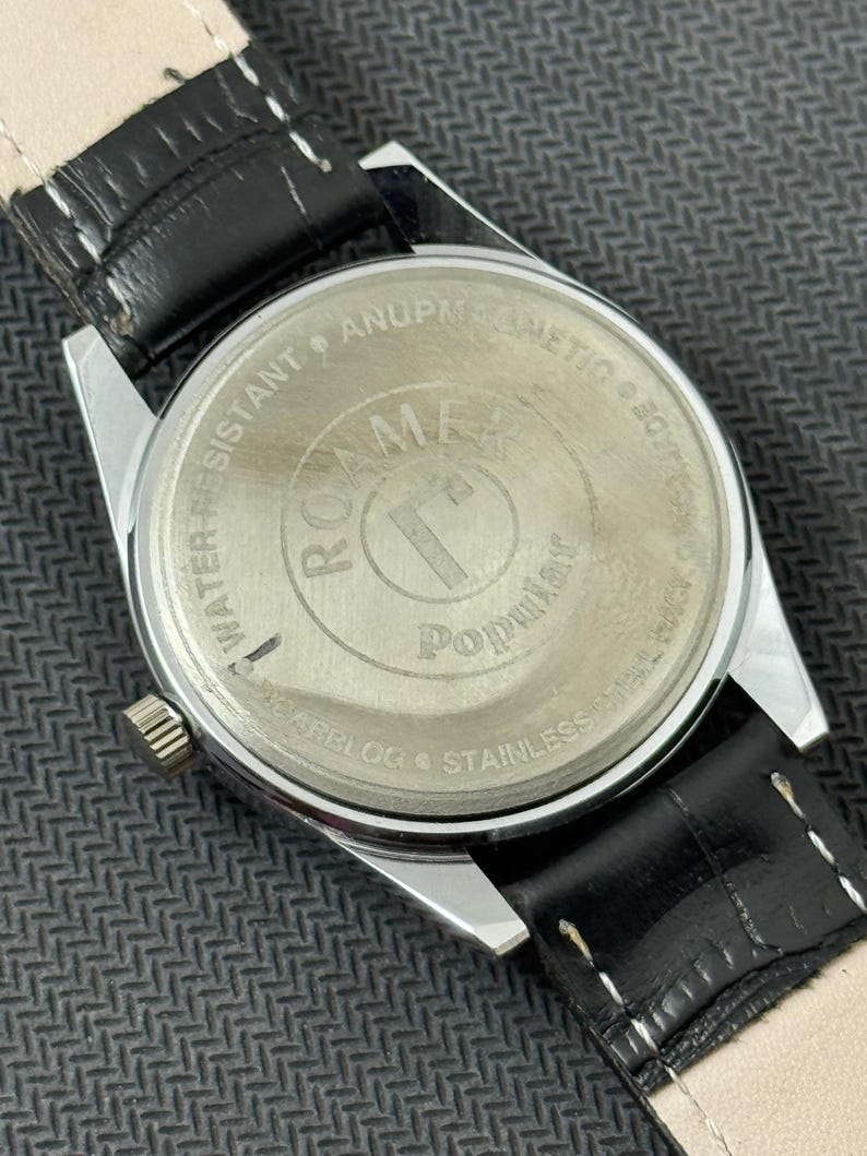 K&ouml;nnte beinhalten: Nahaufnahme der R&uuml;ckseite einer silbernen Uhr mit einem schwarzen Lederarmband. Das Zifferblatt der Uhr tr&auml;gt die Aufschrift "Roamer" und "Popular". Die R&uuml;ckseite der Uhr tr&auml;gt die Aufschrift "Water-Resistant Anuplastic etc. 0.5 ATM Swiss Made Steel Back Stainless."