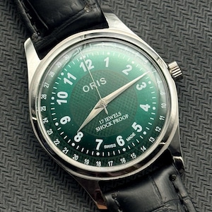 Puede incluir: Un reloj de pulsera plateado con esfera verde y correa de cuero negro. La esfera del reloj muestra la marca "ORIS", números blancos y el texto "17 JEWELS SHOCK PROOF SWISS MADE". El reloj tiene un indicador de fecha.
