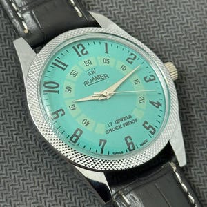 K&ouml;nnte beinhalten: Eine silberne Roamer Armbanduhr mit gr&uuml;nem Zifferblatt und schwarzem Lederband. Das Zifferblatt der Uhr hat wei&szlig;e Markierungen und den Text "RW Roamer" und "17 Jewels Shock Proof".