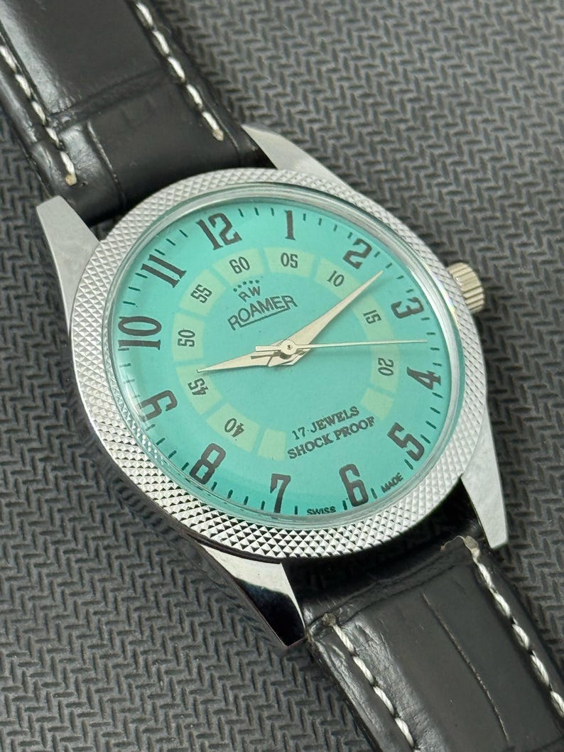 K&ouml;nnte beinhalten: Eine silberne Roamer Armbanduhr mit gr&uuml;nem Zifferblatt und schwarzem Lederband. Das Zifferblatt der Uhr hat wei&szlig;e Markierungen und den Text "RW Roamer" und "17 Jewels Shock Proof".