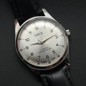 Peut inclure: Une montre ORIS argentée avec un bracelet en cuir noir. Le cadran de la montre est argenté avec des chiffres et des aiguilles noirs. Le texte "ORIS", "17 JEWELS", "SHOCK PROOF" et "SWISS MADE" est visible.