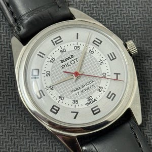 Puede incluir: Un reloj de pulsera plateado con una esfera blanca y una correa de cuero negra. La esfera del reloj tiene marcas negras y una aguja de segundos roja. La esfera del reloj tambi&eacute;n tiene el texto "hmt PILOT" y "PARA SHOCK 17 JEWELS" impreso en ella.