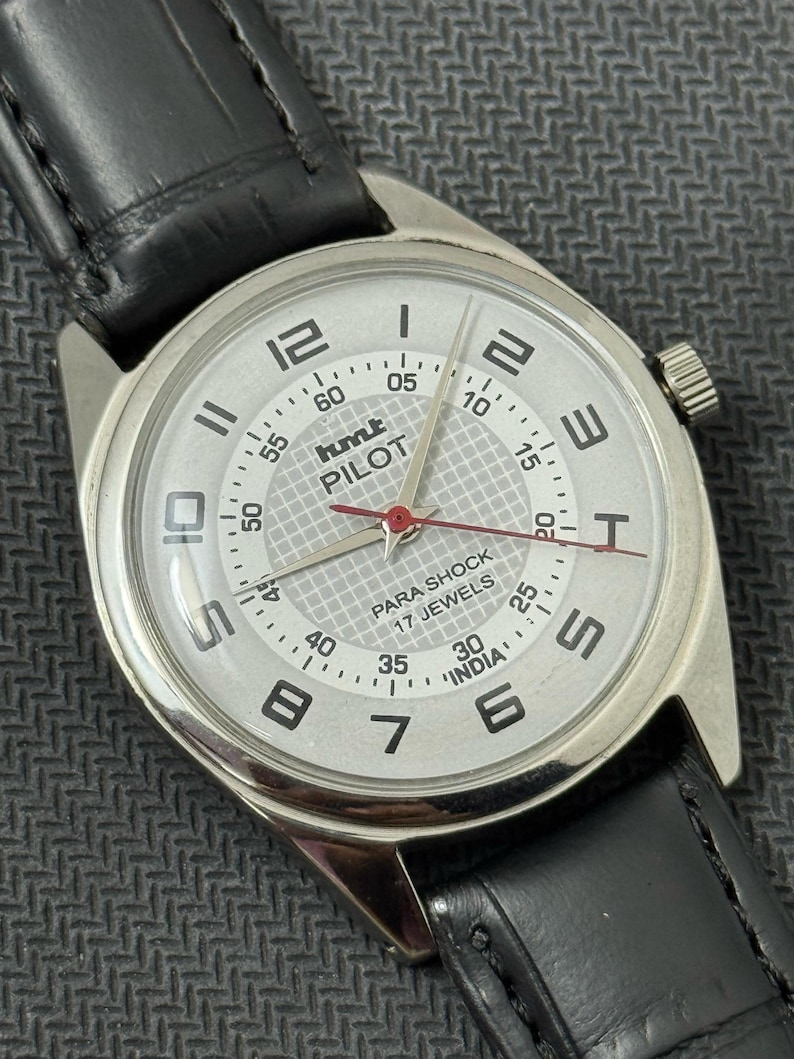 Puede incluir: Un reloj de pulsera plateado con una esfera blanca y una correa de cuero negra. La esfera del reloj tiene marcas negras y una aguja de segundos roja. La esfera del reloj tambi&eacute;n tiene el texto "hmt PILOT" y "PARA SHOCK 17 JEWELS" impreso en ella.