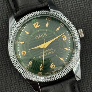 Puede incluir: Un reloj de pulsera Oris vintage con esfera verde y agujas e indicadores dorados. El reloj tiene un bisel de acero inoxidable y una correa de cuero negro. La esfera indica "17 JEWELS SHOCK PROOF" y "SWISS MADE".