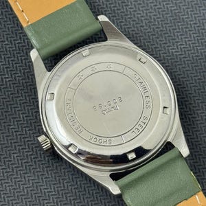 Pode incluir: Um rel&oacute;gio de pulso vintage com caixa de a&ccedil;o inoxid&aacute;vel e pulseira de couro verde. A parte de tr&aacute;s do rel&oacute;gio mostra "Stainless Steel", "Shock Resistant" e o n&uacute;mero "200183". A pulseira tem cerca de 1,9 cm de largura.