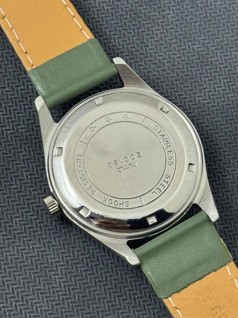Pode incluir: Um rel&oacute;gio de pulso vintage com caixa de a&ccedil;o inoxid&aacute;vel e pulseira de couro verde. A parte de tr&aacute;s do rel&oacute;gio mostra "Stainless Steel", "Shock Resistant" e o n&uacute;mero "200183". A pulseira tem cerca de 1,9 cm de largura.