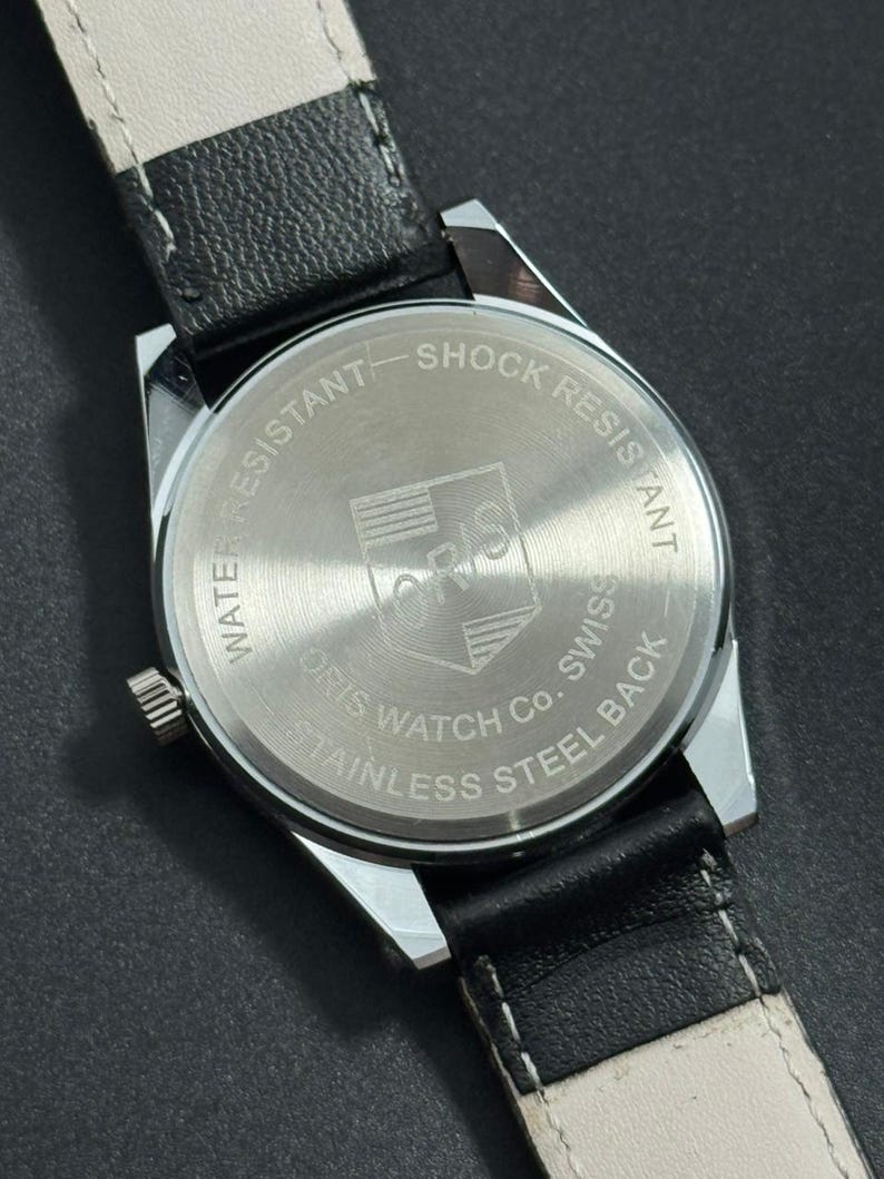 Puede incluir: Primer plano de la parte trasera de un reloj Oris plateado con una correa de cuero negro. La esfera del reloj indica "Water Resistant", "Shock Resistant" y "Stainless Steel Back". El logotipo de Oris es visible en el centro.