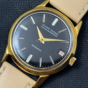 May include: A gold-tone wristwatch with a black face and white markings. The watch face has the text "ULYSSE NARDIN LOCLE & GENEVE" and "AUTOMATIC". The watch has a date window at the 3 o'clock position and a white leather band.