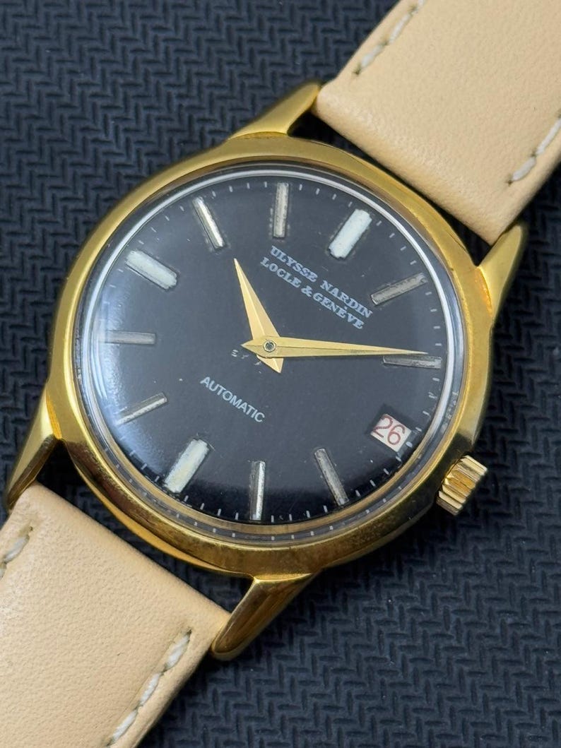 May include: A gold-tone wristwatch with a black face and white markings. The watch face has the text "ULYSSE NARDIN LOCLE & GENEVE" and "AUTOMATIC". The watch has a date window at the 3 o'clock position and a white leather band.