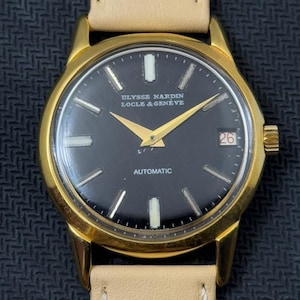 May include: A gold-tone wristwatch with a black face and white markings. The watch face reads "ULYSSE NARDIN LOCLE & GENEVE" and "AUTOMATIC". The date window shows the number 26.