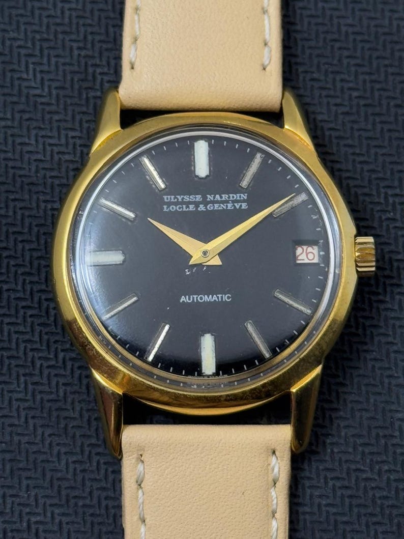 May include: A gold-tone wristwatch with a black face and white markings. The watch face reads "ULYSSE NARDIN LOCLE & GENEVE" and "AUTOMATIC". The date window shows the number 26.