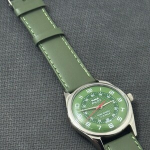 Pode incluir: Rel&oacute;gio anal&oacute;gico verde com caixa prateada e bracelete de couro verde. O mostrador do rel&oacute;gio &eacute; verde com detalhes em branco e vermelho, e o texto "HMT PILOT" &eacute; vis&iacute;vel. A bracelete tem cerca de 2 cm de largura.