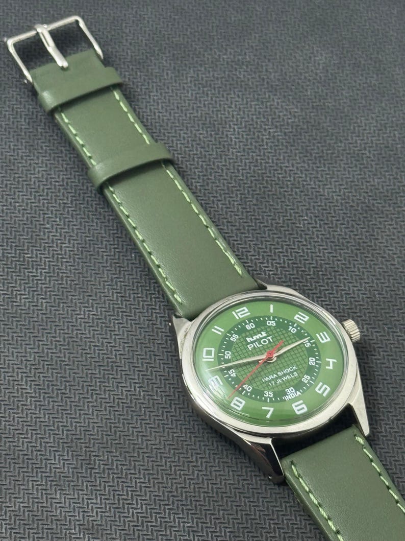 Pode incluir: Rel&oacute;gio anal&oacute;gico verde com caixa prateada e bracelete de couro verde. O mostrador do rel&oacute;gio &eacute; verde com detalhes em branco e vermelho, e o texto "HMT PILOT" &eacute; vis&iacute;vel. A bracelete tem cerca de 2 cm de largura.