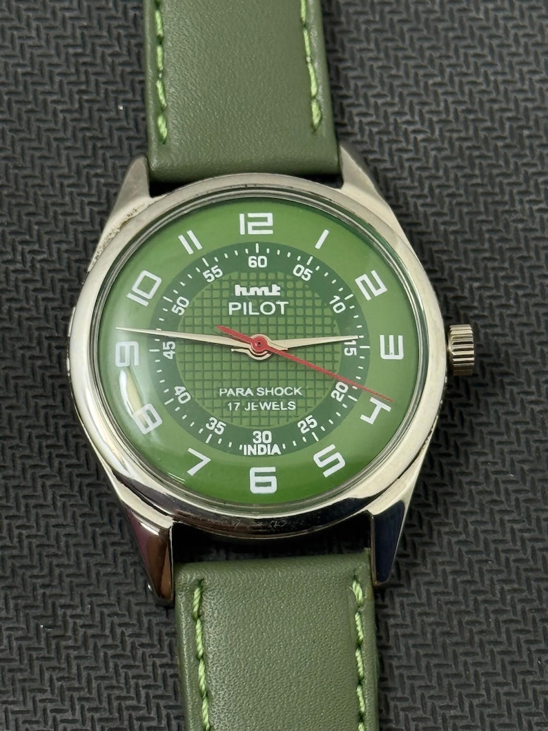 Pode incluir: Um rel&oacute;gio de pulso verde com caixa prateada e pulseira de couro verde. O mostrador do rel&oacute;gio &eacute; verde com algarismos e ponteiros brancos, e o texto "PILOT" e "INDIA" s&atilde;o vis&iacute;veis.