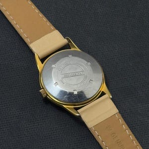 May include: A gold-tone wristwatch with a tan leather band. The back of the watch has the words "WATERPROOF" and "SWISS MADE" printed on it.