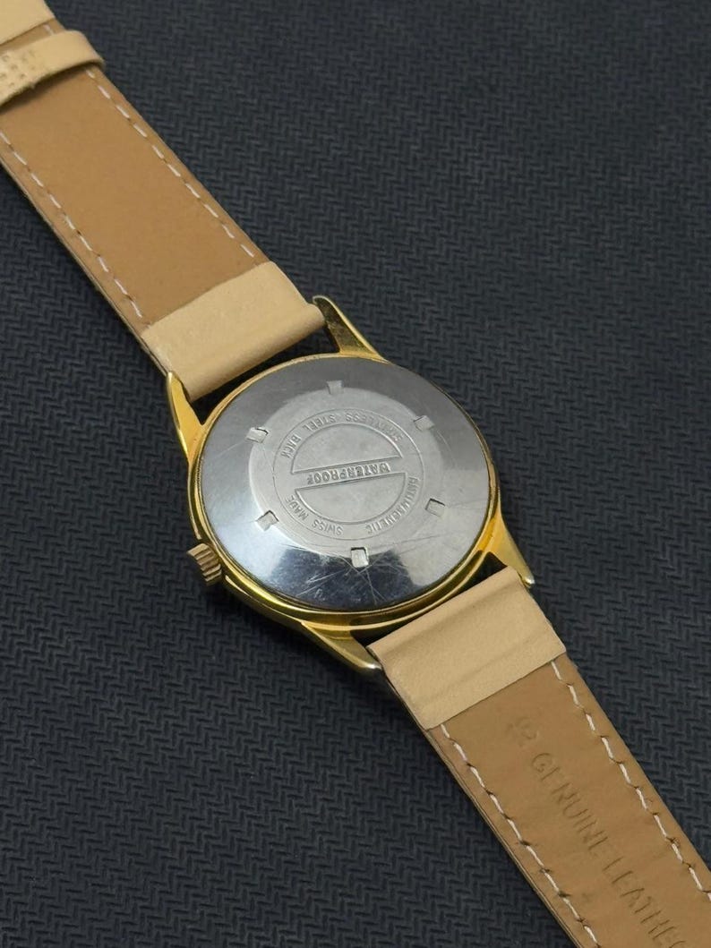 May include: A gold-tone wristwatch with a tan leather band. The back of the watch has the words "WATERPROOF" and "SWISS MADE" printed on it.