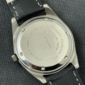 Puede incluir: Un reloj plateado con una correa de cuero negro. La parte posterior del reloj tiene el texto "STAINLESS STEEL - WATER RESISTANT - SHOCK RESISTANT - MADE IN INDIA - 051187".