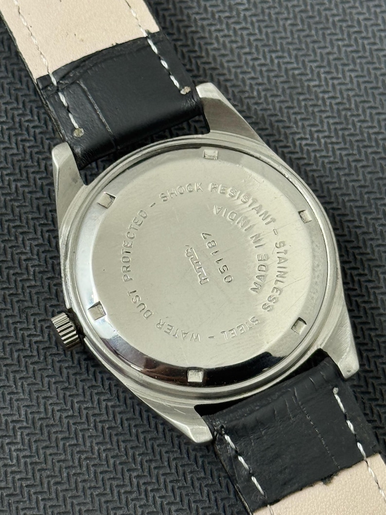 Puede incluir: Un reloj plateado con una correa de cuero negro. La parte posterior del reloj tiene el texto "STAINLESS STEEL - WATER RESISTANT - SHOCK RESISTANT - MADE IN INDIA - 051187".