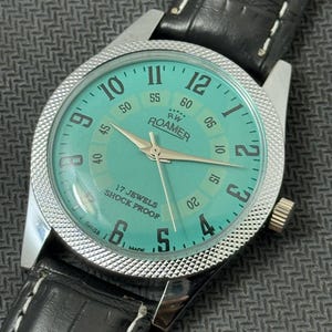 K&ouml;nnte beinhalten: Eine silberne Roamer Armbanduhr mit gr&uuml;nem Zifferblatt und schwarzem Lederband. Das Zifferblatt der Uhr hat wei&szlig;e Ziffern und Zeiger, und die Worte "RW Roamer" und "17 Jewels Shock Proof" sind auf dem Zifferblatt aufgedruckt.