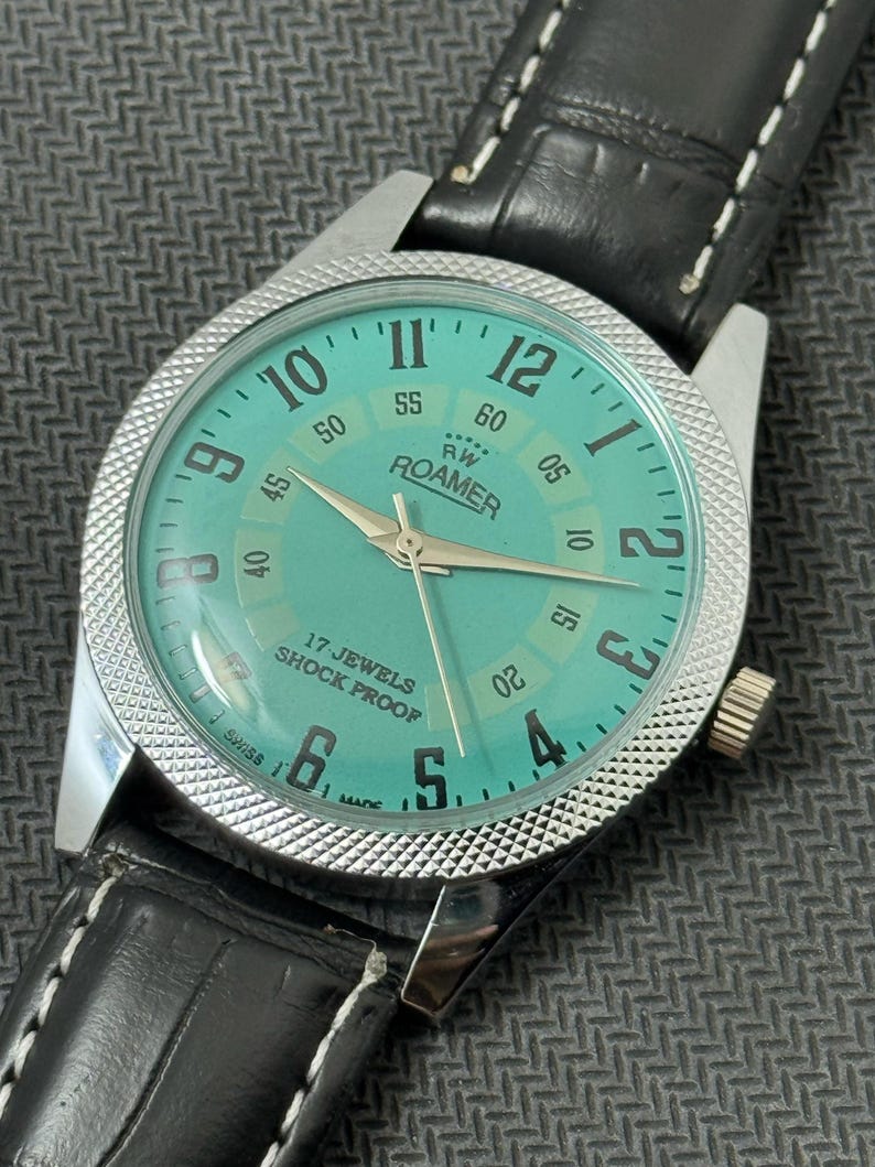 K&ouml;nnte beinhalten: Eine silberne Roamer Armbanduhr mit gr&uuml;nem Zifferblatt und schwarzem Lederband. Das Zifferblatt der Uhr hat wei&szlig;e Ziffern und Zeiger, und die Worte "RW Roamer" und "17 Jewels Shock Proof" sind auf dem Zifferblatt aufgedruckt.