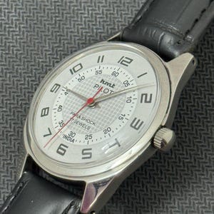 Puede incluir: Un reloj de pulsera plateado con una esfera blanca y una correa de cuero negra. La esfera del reloj tiene n&uacute;meros y marcas negras, una aguja de segundos roja y las palabras "hmt PILOT" impresas en ella. El reloj tambi&eacute;n tiene las palabras "PARA SHOCK 17 JEWELS INDIA" impresas en la esfera.