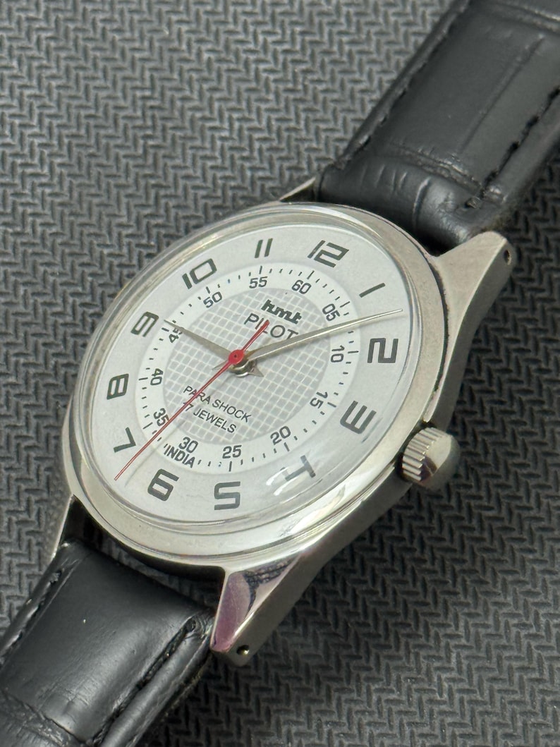 Puede incluir: Un reloj de pulsera plateado con una esfera blanca y una correa de cuero negra. La esfera del reloj tiene n&uacute;meros y marcas negras, una aguja de segundos roja y las palabras "hmt PILOT" impresas en ella. El reloj tambi&eacute;n tiene las palabras "PARA SHOCK 17 JEWELS INDIA" impresas en la esfera.