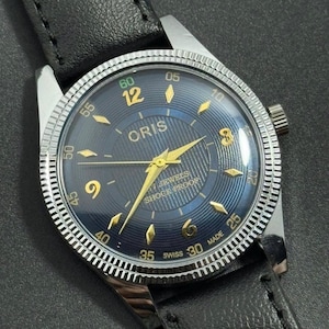 Puede incluir: Un reloj de pulsera Oris vintage con esfera azul oscuro, agujas y n&uacute;meros dorados, y bisel plateado. El reloj tiene una correa de cuero negro y est&aacute; marcado "17 JEWELS SHOCK PROOF". La esfera mide aproximadamente 3,5 cm de di&aacute;metro.