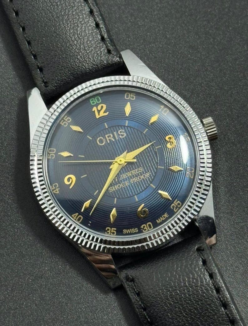 Puede incluir: Un reloj de pulsera Oris vintage con esfera azul oscuro, agujas y n&uacute;meros dorados, y bisel plateado. El reloj tiene una correa de cuero negro y est&aacute; marcado "17 JEWELS SHOCK PROOF". La esfera mide aproximadamente 3,5 cm de di&aacute;metro.