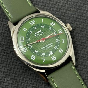Pode incluir: Um rel&oacute;gio de pulso com mostrador verde, caixa prateada e pulseira de couro verde. O mostrador apresenta algarismos e ponteiros brancos, com o texto "PILOT" e "PARA SHOCK 17 JEWELS". A pulseira &eacute; costurada com linha verde clara.