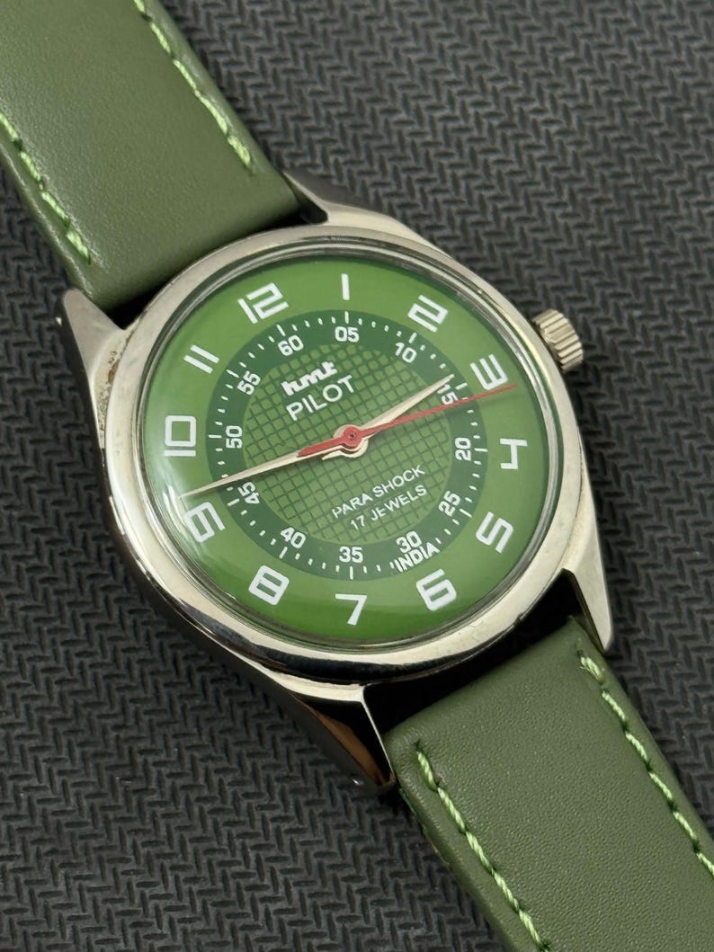 Pode incluir: Um rel&oacute;gio de pulso com mostrador verde, caixa prateada e pulseira de couro verde. O mostrador apresenta algarismos e ponteiros brancos, com o texto "PILOT" e "PARA SHOCK 17 JEWELS". A pulseira &eacute; costurada com linha verde clara.
