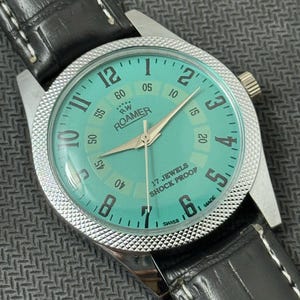 K&ouml;nnte beinhalten: Eine silberfarbene Armbanduhr mit einem t&uuml;rkisfarbenen Zifferblatt. Das Zifferblatt der Uhr hat schwarze r&ouml;mische Ziffern f&uuml;r die Stunden und schwarze Minutenmarkierungen am &auml;u&szlig;eren Rand. Das Zifferblatt der Uhr hat auch ein wei&szlig;es Zifferblatt mit schwarzem Text, der "RW ROAMER" und "17 JEWELS SHOCK PROOF" lautet. Die Uhr hat ein schwarzes Lederarmband mit wei&szlig;en N&auml;hten.