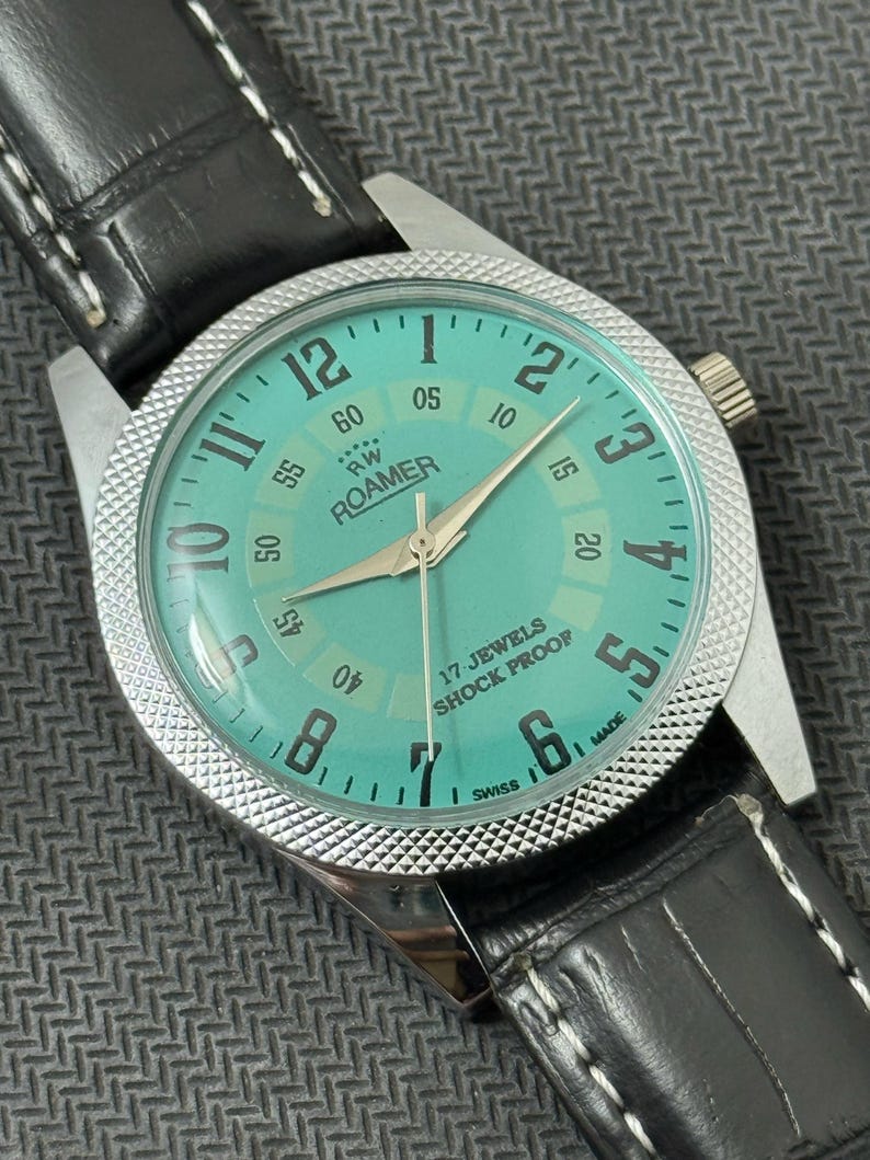 K&ouml;nnte beinhalten: Eine silberfarbene Armbanduhr mit einem t&uuml;rkisfarbenen Zifferblatt. Das Zifferblatt der Uhr hat schwarze r&ouml;mische Ziffern f&uuml;r die Stunden und schwarze Minutenmarkierungen am &auml;u&szlig;eren Rand. Das Zifferblatt der Uhr hat auch ein wei&szlig;es Zifferblatt mit schwarzem Text, der "RW ROAMER" und "17 JEWELS SHOCK PROOF" lautet. Die Uhr hat ein schwarzes Lederarmband mit wei&szlig;en N&auml;hten.