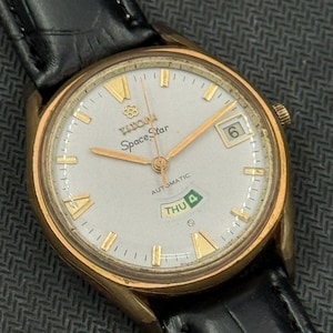 Op de afbeelding: Vintage Titan Space Star automatische polshorloge met een goudkleurige kast en een witte wijzerplaat. Het horloge heeft gouden wijzers en markeringen, een dag-datumweergave en een zwarte leren band. De wijzerplaat leest "Space Star" en "Automatic".
