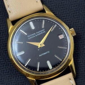 May include: A gold-tone wristwatch with a black face and white markings. The watch face says "ULYSSE NARDIN LOCLE & GENEVE" and "AUTOMATIC". The date window shows the number "26".