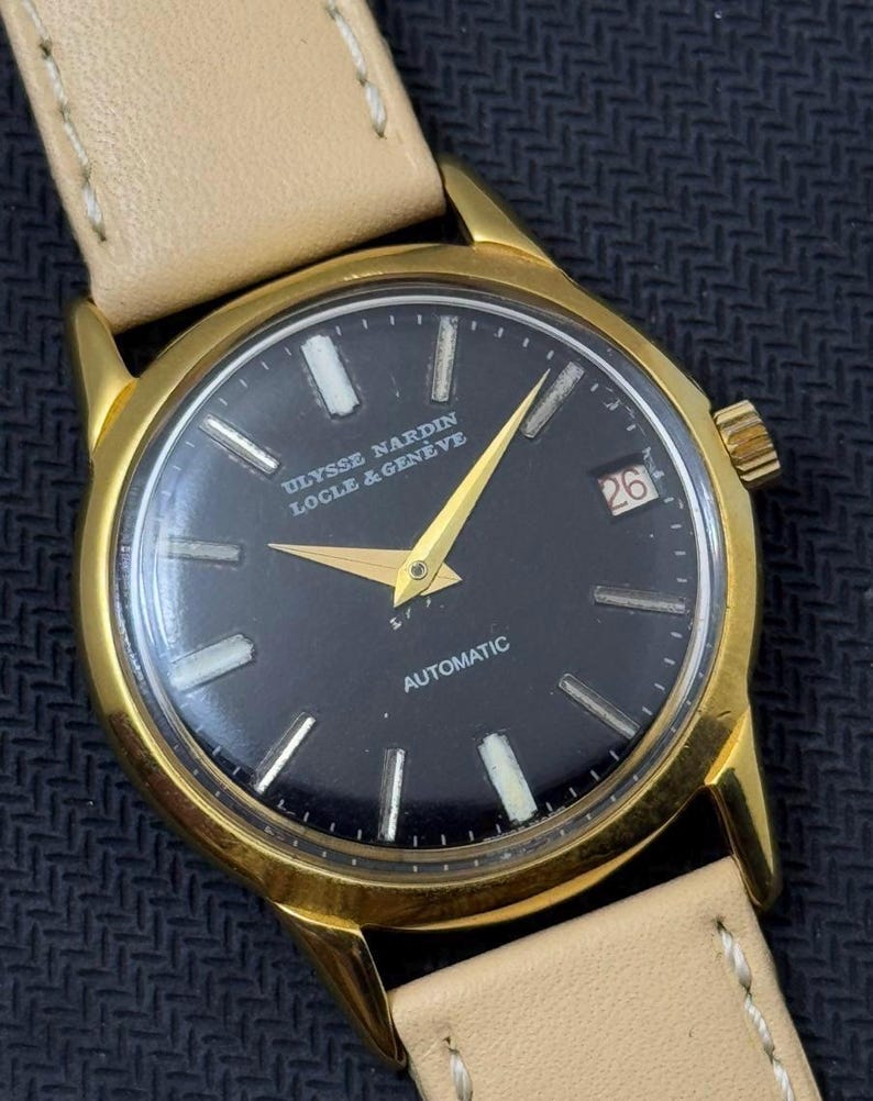May include: A gold-tone wristwatch with a black face and white markings. The watch face says "ULYSSE NARDIN LOCLE & GENEVE" and "AUTOMATIC". The date window shows the number "26".