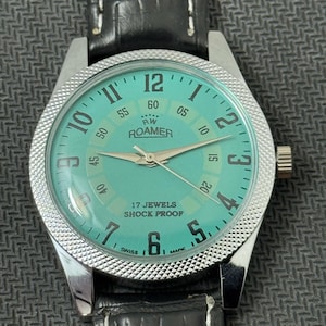 K&ouml;nnte beinhalten: Eine silberfarbene Armbanduhr mit einem schwarzen Lederarmband. Das Zifferblatt der Uhr ist hellblau mit schwarzen Markierungen und den Worten "RW ROAMER" und "17 JEWELS SHOCK PROOF".