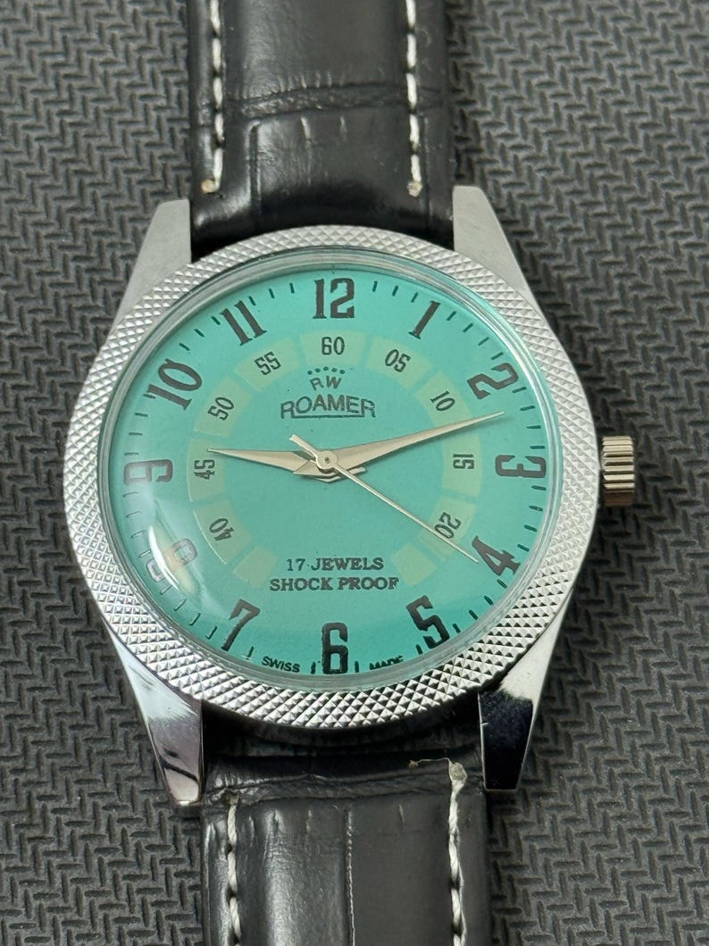 K&ouml;nnte beinhalten: Eine silberfarbene Armbanduhr mit einem schwarzen Lederarmband. Das Zifferblatt der Uhr ist hellblau mit schwarzen Markierungen und den Worten "RW ROAMER" und "17 JEWELS SHOCK PROOF".
