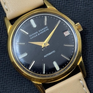 May include: A gold-tone wristwatch with a black face and white markings. The watch has a date window at the 3 o'clock position and the text "ULYSSE NARDIN LOCLE & GENEVE" and "AUTOMATIC" printed on the face.