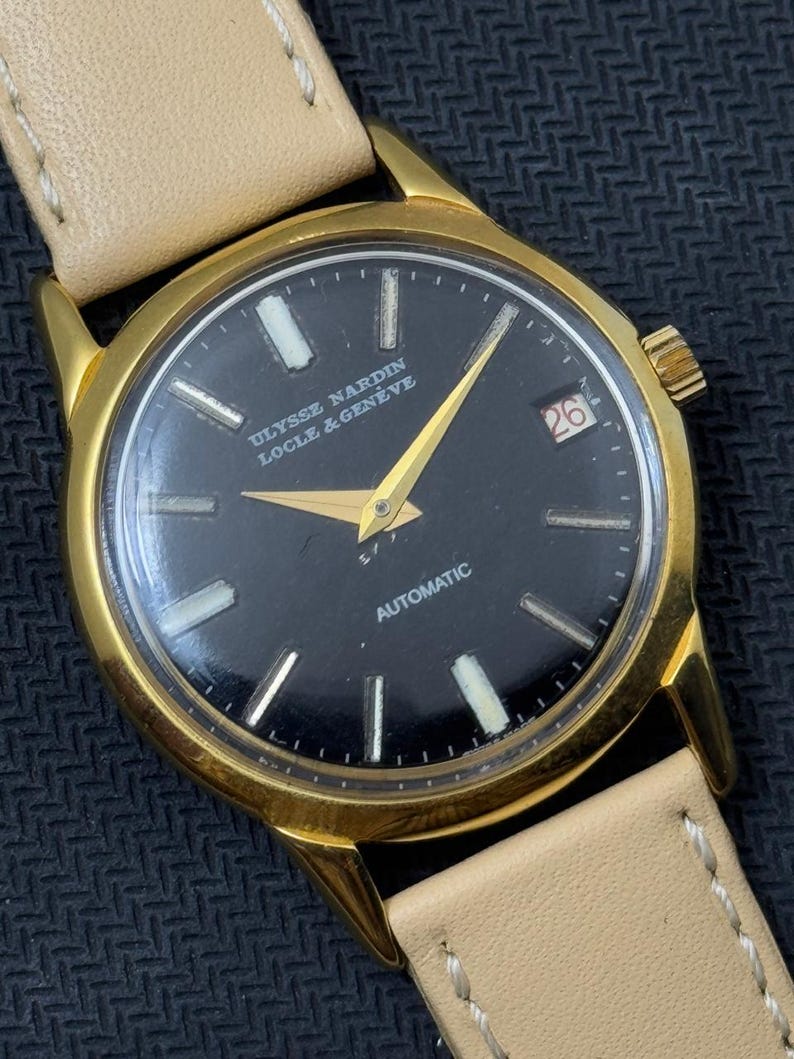 May include: A gold-tone wristwatch with a black face and white markings. The watch has a date window at the 3 o'clock position and the text "ULYSSE NARDIN LOCLE & GENEVE" and "AUTOMATIC" printed on the face.