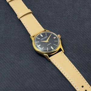 May include: A gold-tone wristwatch with a black face and a tan leather band. The watch face has the words "Dugena-Werke" and "Automatic" printed on it.