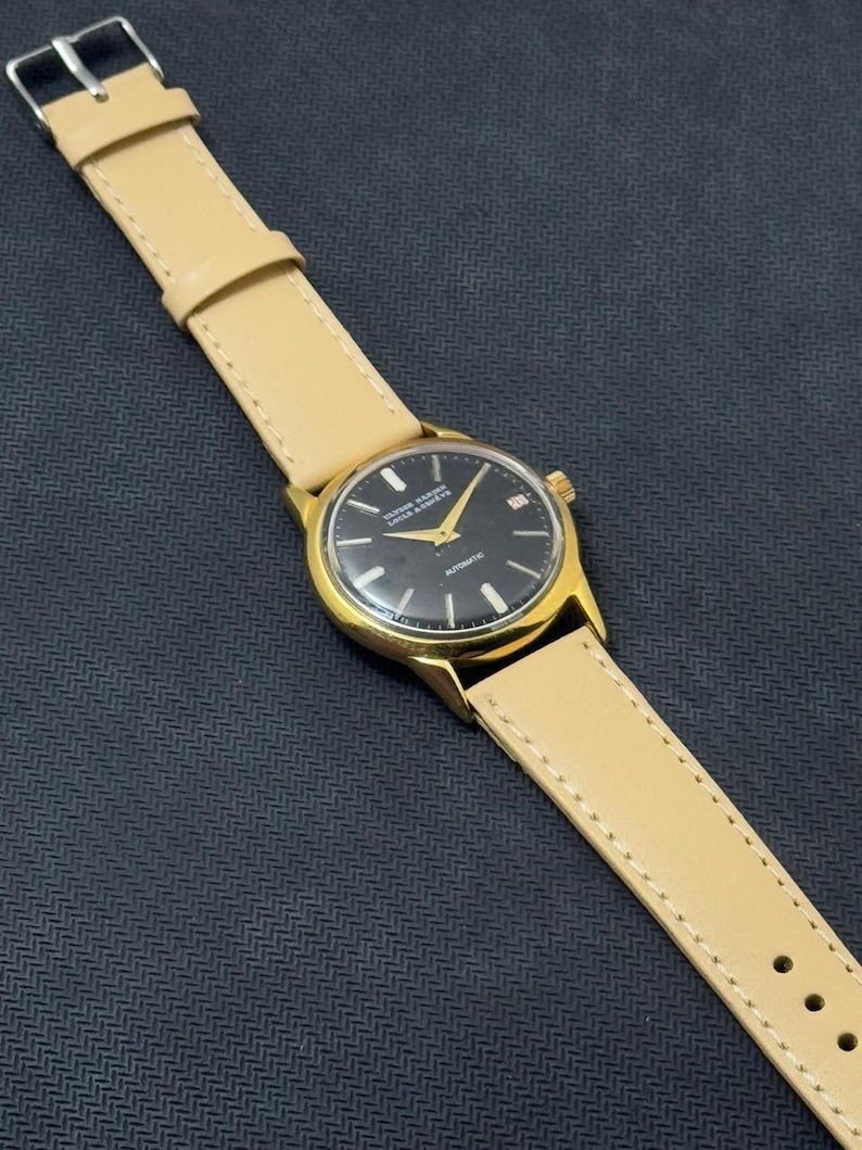 May include: A gold-tone wristwatch with a black face and a tan leather band. The watch face has the words "Dugena-Werke" and "Automatic" printed on it.