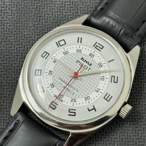 Puede incluir: Un reloj de pulsera plateado con una esfera blanca y una correa de cuero negra. La esfera del reloj tiene marcas negras y el texto "hmt PILOT PARA SHOCK 17 JEWELS INDIA".