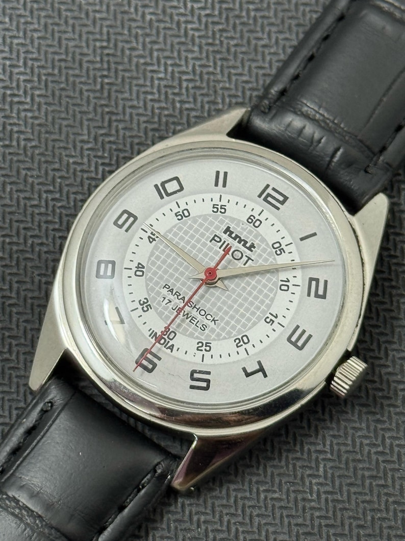 Puede incluir: Un reloj de pulsera plateado con una esfera blanca y una correa de cuero negra. La esfera del reloj tiene marcas negras y el texto "hmt PILOT PARA SHOCK 17 JEWELS INDIA".