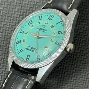 K&ouml;nnte beinhalten: Eine silberne Roamer Armbanduhr mit einem schwarzen Lederarmband und einem t&uuml;rkisblauen Zifferblatt. Das Zifferblatt hat wei&szlig;e Markierungen f&uuml;r die Zahlen und einen wei&szlig;en Sekundenzeiger. Das Zifferblatt der Uhr tr&auml;gt au&szlig;erdem den Text "RW Roamer" und "17 Jewels Shock Proof".